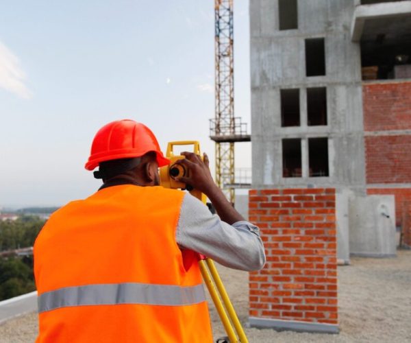 African American young man topographer in casque measuring angle with total station on roof of building. Male builder. Constructor doing topographic measures. Geodesy concept. Geodesic. Constructing.